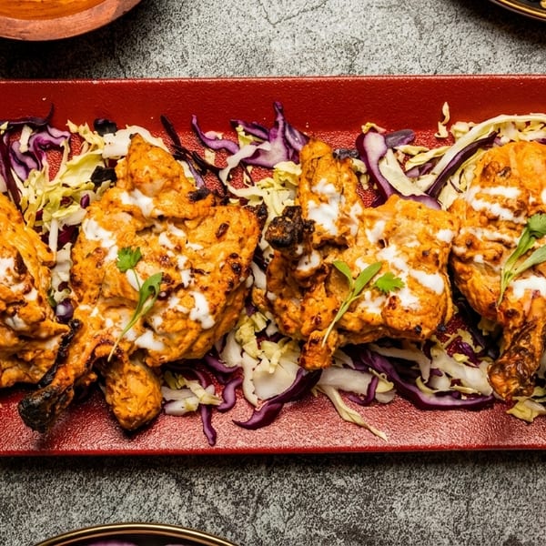Four pieces of tandoori chicken with white sauce, cabbage, and a lime wedge on a red plate.