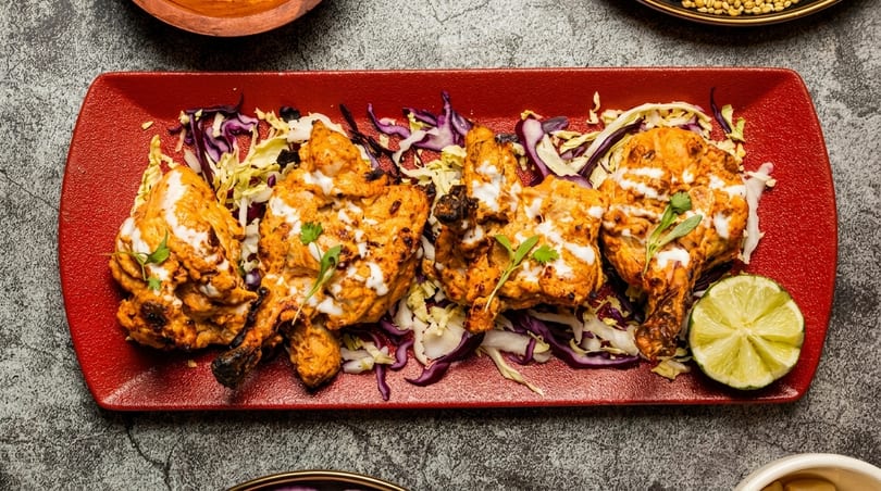 Four pieces of tandoori chicken with white sauce, cabbage, and a lime wedge on a red plate.