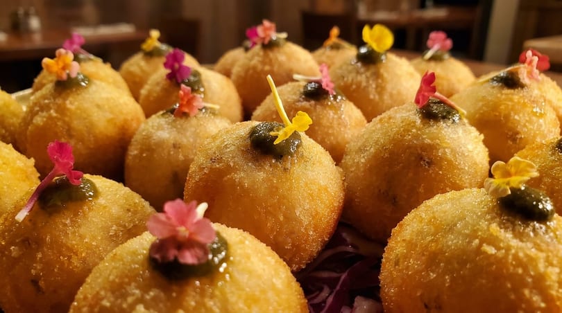 Golden fried arancini balls, each topped with green sauce and a small colorful edible flower.