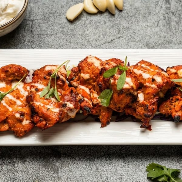 Grilled chicken tikka with white sauce drizzle, lime, garlic, and spices on a white plate.