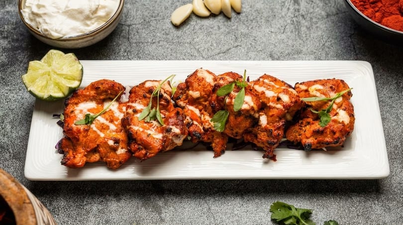 Grilled chicken tikka with white sauce drizzle, lime, garlic, and spices on a white plate.