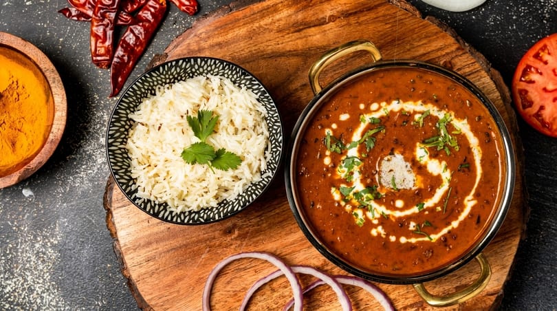 A bowl of white rice with cilantro and a pot of creamy dal makhani on a wooden board.