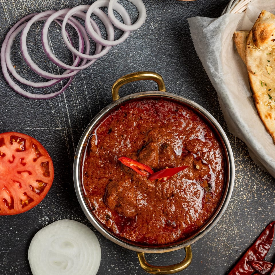 Rich red curry in a metal bowl, with naan, onions, tomato, and spices on a dark surface.