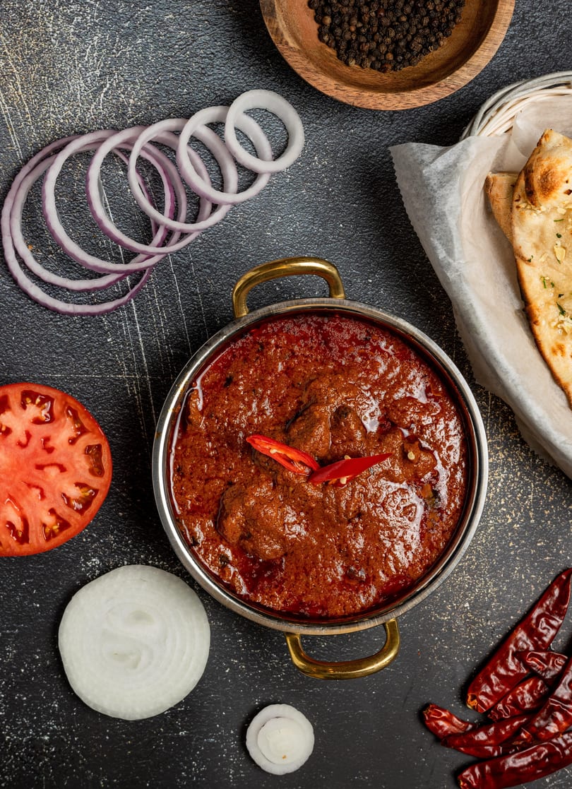 Rich red curry in a metal bowl, with naan, onions, tomato, and spices on a dark surface.