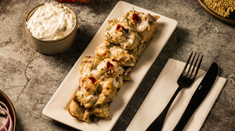 Creamy chicken tikka on a white platter with a side of dipping sauce and black cutlery.