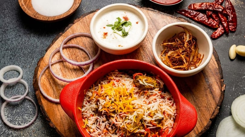Biryani in a red pot with raita, fried onions, and fresh onion rings on a wooden board.