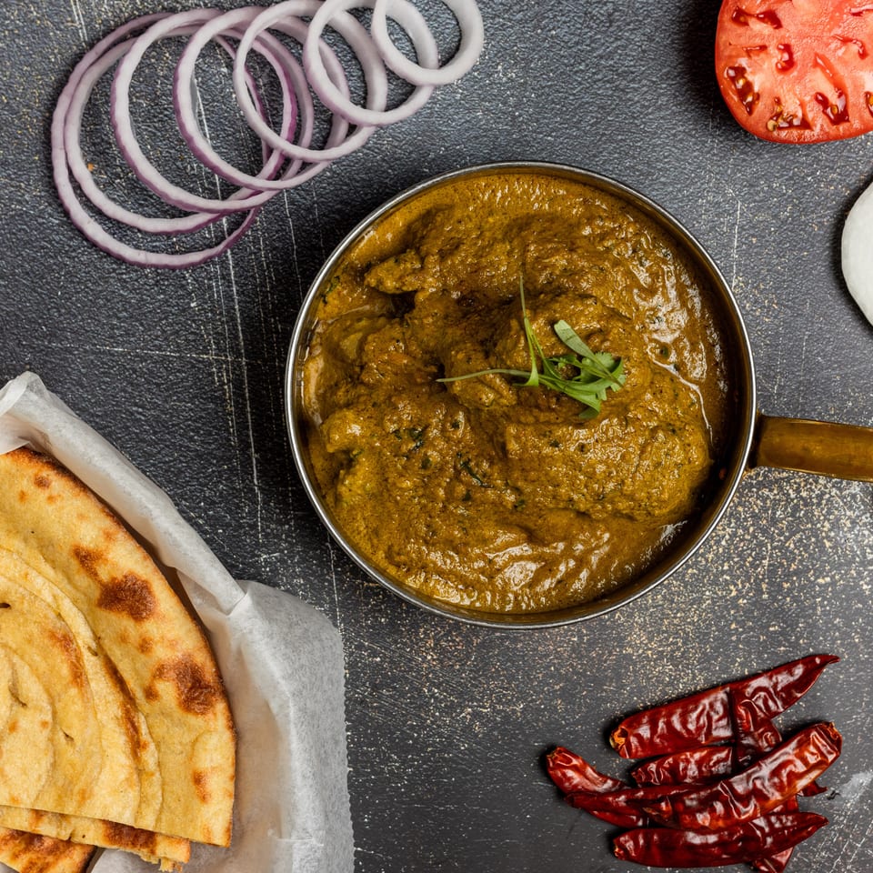 Overhead view of Indian curry in a brass pot, with naan bread, red onion, tomato, and dried chilies.
