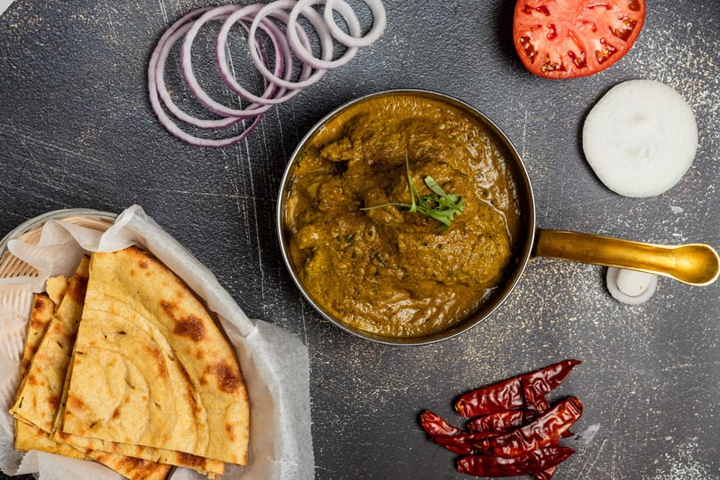 Overhead view of Indian curry in a brass pot, with naan bread, red onion, tomato, and dried chilies.