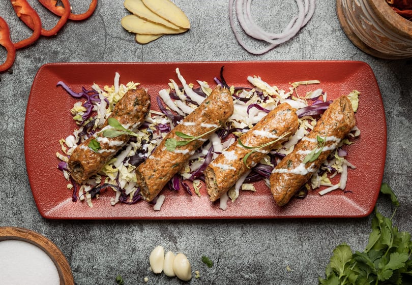 Four kebabs with white sauce and herbs on a bed of shredded cabbage, on a red rectangular plate.