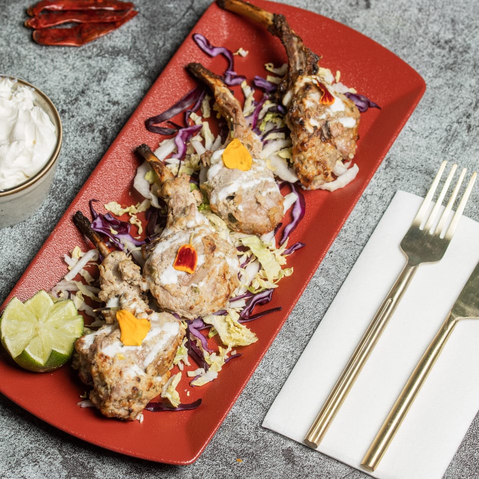 Four lamb chops on a red plate with shredded cabbage, a lime wedge, and a side of white sauce.