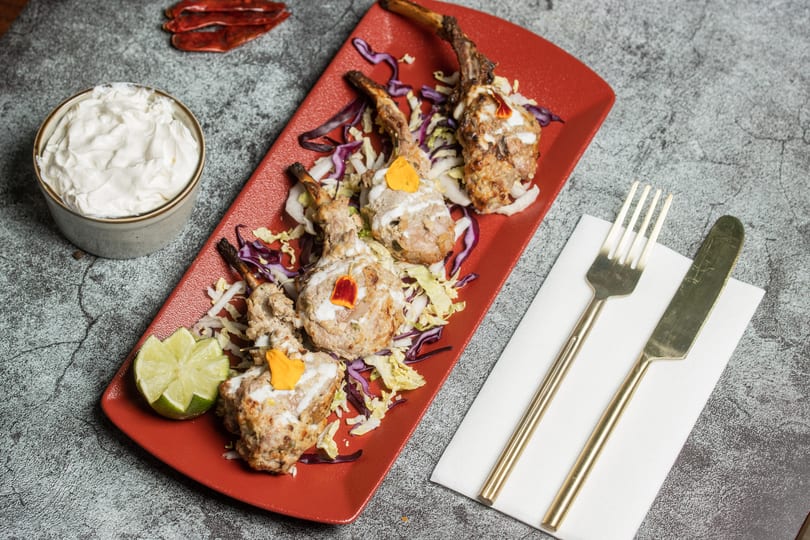 Four lamb chops on a red plate with shredded cabbage, a lime wedge, and a side of white sauce.
