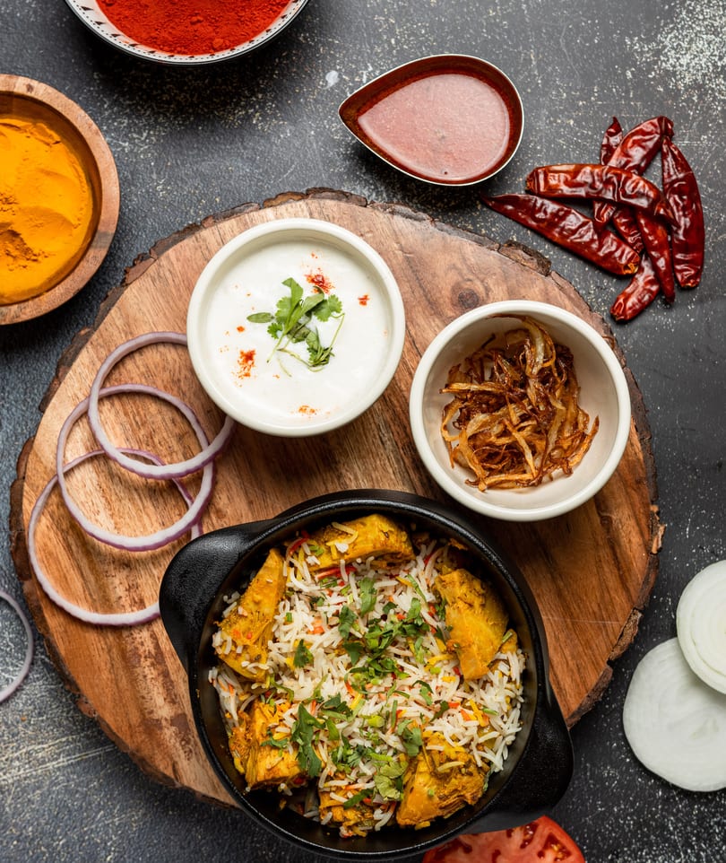 Chicken biryani in a black pot, with sides of yogurt, fried onions, and spices on a wooden board.