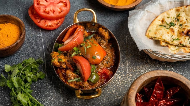 Indian curry with tomatoes and cilantro in a brass pot, with naan bread and spices.